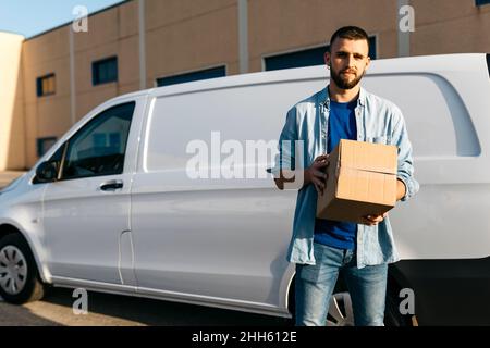 Young delivery man with package standing in front of van Stock Photo
