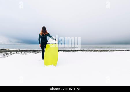 Man with surfboard standing on snow covered beach Stock Photo