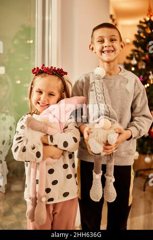 Happy sibling with toy gnomes standing in front of christmas tree at home Stock Photo