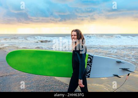 Smiling surfer in wetsuit walking with surfboard near sea