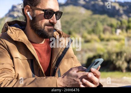 Bearded man wearing sunglasses using mobile phone and listening music through wireless in-ear headphones Stock Photo