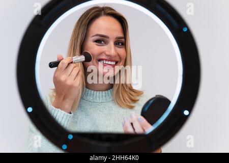 Young woman applying blusher in front of ring light Stock Photo