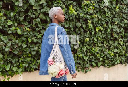 Woman walking by plants carrying groceries in mesh bag Stock Photo