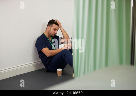 Worried male nurse with head in hand sitting by wall in medical room Stock Photo