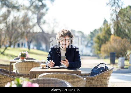 Man using smart phone writing in diary at sidewalk cafe on sunny day Stock Photo