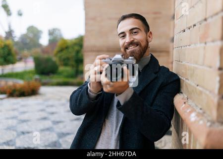 Happy young bearded man with camera leaning on wall Stock Photo