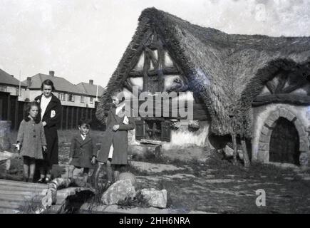 1930s, historical, in a suburban area, young children standing outside a strange looking, 'hobbit' type minature or toy house, England, UK. With its uneven curved timbers, tiny windows and doors and a sloping grass covered roof, this quirky dwelling is an unusual building and possibly, even in this era, was a visitor attraction. Stock Photo