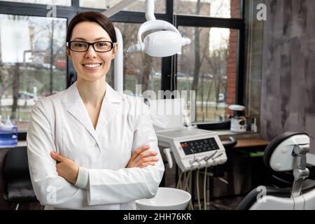 Portrait of a young female dentist on the background of the clinic. The dentist is standing with his arms crossed, looking at the camera and smiling. Stock Photo