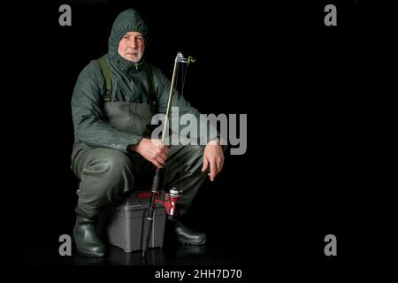A fisherman in waders and raincoat sits on his fishing box against a black background and holds a fishing rod in his hand. Stock Photo
