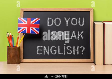 a chalkboard with the text do you speak english? written in it, a pot with pencils and the flag of the United Kingdom, on a wooden desk, with green ba Stock Photo