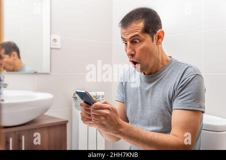 Latin man receiving surprising news on line in a smart phone at the bathroom. Stock Photo