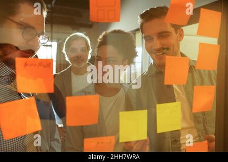 Diverse group of businesspeople laughing while standing in an office brainstorming together with sticky notes on a glass wall Stock Photo