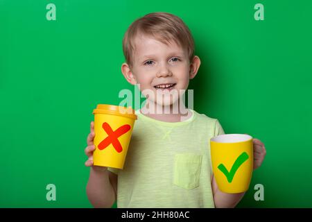 Ecology concept. Child kid boy with reusable mug and disposable paper cup. Studio shoot on green background. Stock Photo