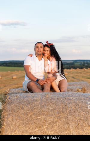 Loving couple in a field on rolls of straw. Young man and woman having fun, hugging and kissing on sunset. Stock Photo