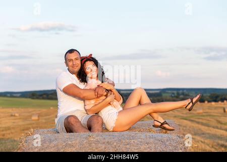 Loving couple in a field on rolls of straw. Young man and woman having fun, hugging and kissing on sunset. Stock Photo