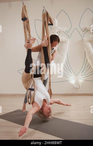 Vertical full length shot of a professional fly yoga trainer helping senior client doing decompression exercise on a hammock Stock Photo