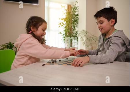 Two adorable kids, boy and girl, brother and sister having great time playing checkers board game together at home interior. Stock Photo