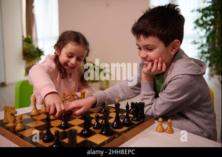 Two adorable kids, boy and girl, brother and sister having great time playing chess game together at home interior. Stock Photo
