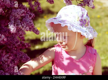 Beautiful little blonde girl with panama on her head playing in spring garden with blooming bush of purple lilac. Stock Photo