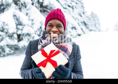 polyethnic happy man in stylish coat and hat holding gift box with red ribbon in winter park, valentine s day concept Stock Photo