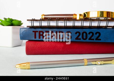 On a wooden table there are reports, a potted plant, a magnifying glass, a black pen and a notebook with the text TRENDS 2022. Business concept Stock Photo