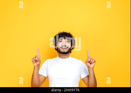 Cheerful curly haired millennial Indian or Arabian guy, pointing fingers up at empty space, looks up, smiling friendly, stands on isolated orange background in casual white t-shirt, mock-up concept Stock Photo