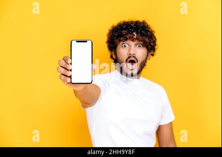 Defocused surprised Indian young man in white t-shirt, holds smartphone with mock-up empty white screen, stands on isolated orange background, shocked looks at the camera Stock Photo