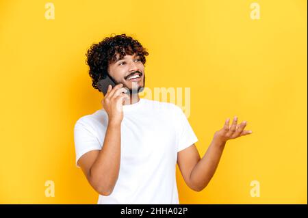 Phone talk by cellphone. Happy joyful excited Indian charismatic guy has a pleasant phone conversation, happy facial expression, stands on isolated orange background, gesturing hand, smiling Stock Photo