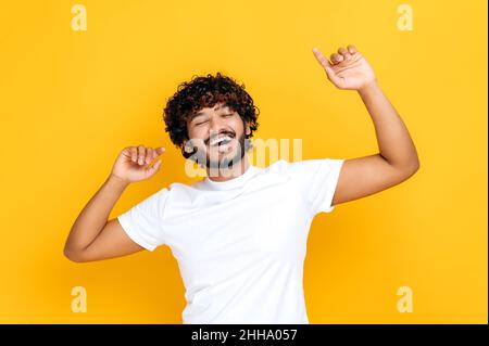 Joy, victory, luck, dancing. Cheerful happy Indian guy in casual white t-shirt, dancing to favorite music, having fun, rejoice in luck, celebrating win, on isolated orange background, smiling Stock Photo