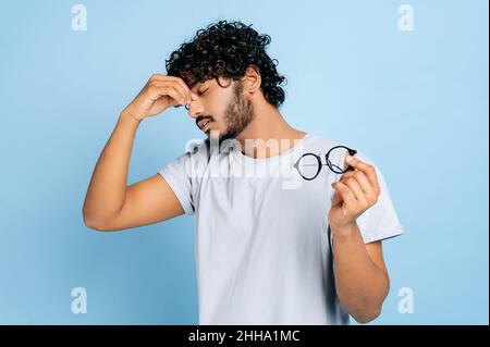 Tired exhausted indian or arab guy in a t-shirt, took off his glasses, closed his eyes, massages the bridge of his nose, has a migraine, headache, stands on an isolated blue background Stock Photo