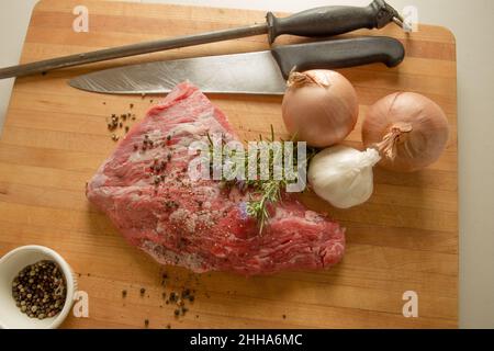 Try tip of Beef on a butchers wood cutting board with fresh rosemary, sage, onion, garlic, pepper and seasoning. chefs knife and sharpening steel. Stock Photo