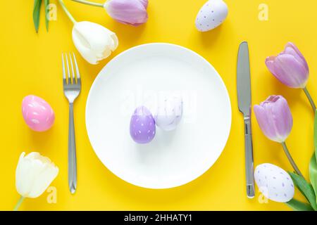 Easter table setting. White plate, decorative easter eggs, tulips and silver cutlery on yellow background Stock Photo