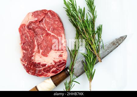 Close-up of a raw rib eye steak, one knife and a few sprigs of fresh rosemary on a white surface. Food concept. Overhead view. Stock Photo