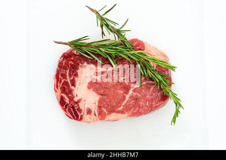 Close-up of a raw rib eye steak and a few sprigs of fresh rosemary on a white surface. Food concept. Overhead view. Stock Photo