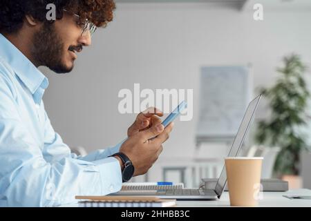 Indian man using mobile phone app sitting front of laptop on desk side view shot Stock Photo