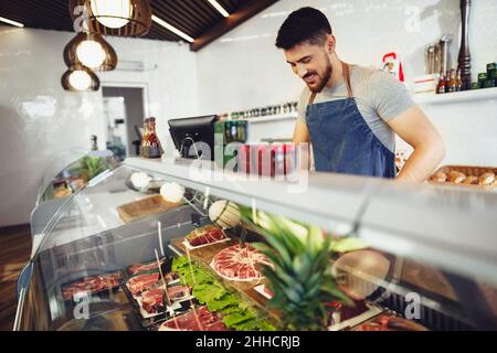 Young man butcher arranging meat products in display case of butcher shop Stock Photo