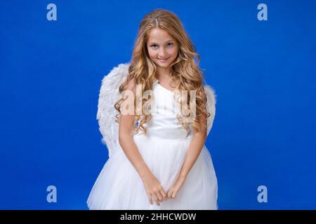 Angel girl with wings, valentine's day. Child with an angelic character. Little girl in an angel costume, white dress and feather wings on a blue background. The concept of an innocent child. Stock Photo