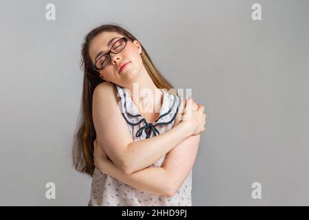 Emotions and people. A young brunette woman in glasses and a blouse, closing her eyes, hugs herself by the shoulders. Emotion of calm. Stock Photo