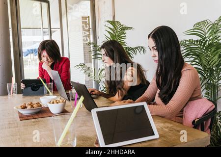 Pensive women in casual clothes browsing data on digital gadgets while sitting at table near snacks and tablet with black screen and working remotely Stock Photo