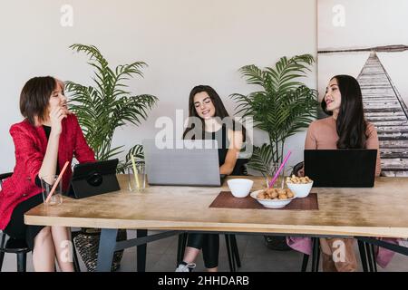 Cheerful female freelancers smiling and talking with each other while sitting at table with digital gadgets and snacks and taking break in remote job Stock Photo