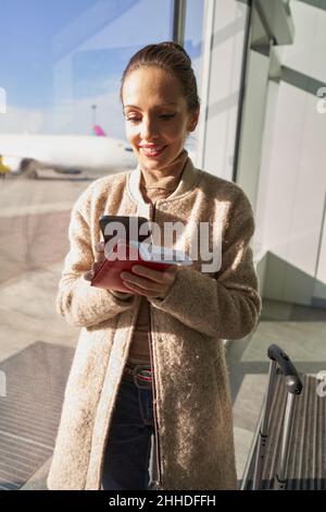 Cheerful lady in coat using smartphone near airport window Stock Photo