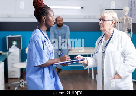 Multi ethnic medical team taking notes on clipboard about healthcare of patient, having discussion about diagnosis and prescription treatment. Doctor and nurse talking about medicine Stock Photo