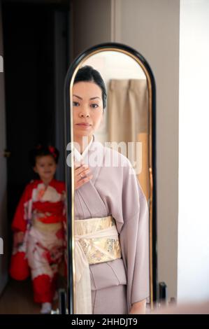 Little girl wearing red kimono watching her mother getting dressed in Japanese traditional clothes in front of the mirror. Stock Photo