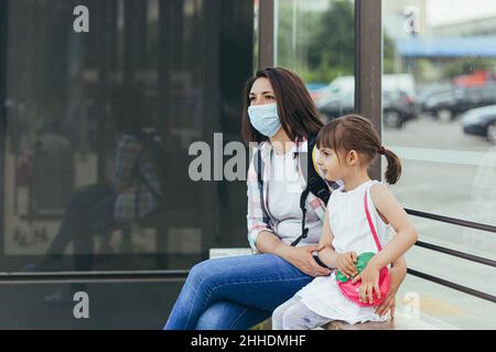 Young woman wearing a protective mask with her daughter waiting for a public bus at the bus station Stock Photo