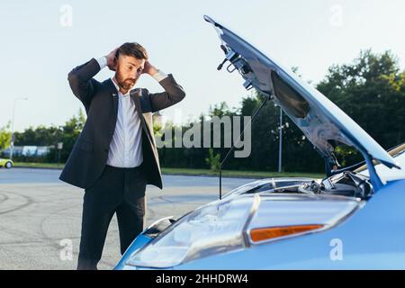 an annoyed and angry man, near a broken car, tries to call an insurance agent and a repair crew. for car repair Stock Photo