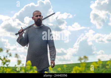 Bald bearded man in  metal chain mail over  linen shirt stands in  middle of  field, holding  sword. Militant look. Blurred background. Stock Photo