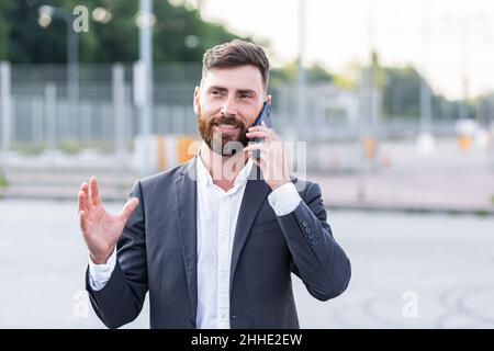 Happy bearded man looks in reverse and talks on the phone, businessman smiles and talks to colleague on the phone, outside the office during lunch bre Stock Photo
