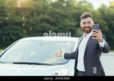 Successful and happy man brags about buying a new car, having fun talking to friends uses a video call in the phone app Stock Photo