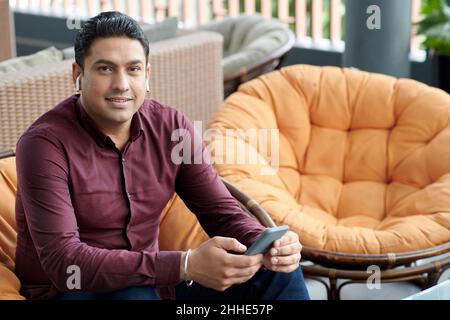 Portrait of smiling businessman in maroon shirt sitting in armchair in lounge area and wearing earbuds when calling coworker Stock Photo