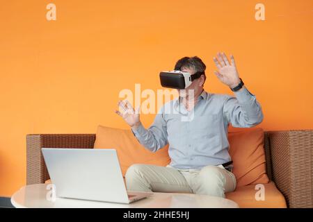 Senior businessman sitting on sofa in virtual reality glasses and testing new application Stock Photo
